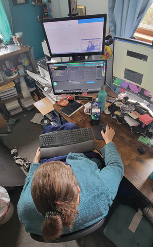 Shawna sitting in front of a cluttered and messy desk, looking at 3 computer monitors.