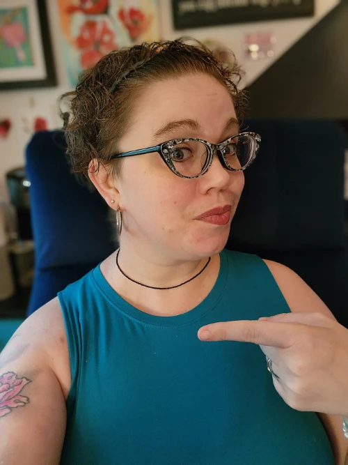 Shawna in a teal sleeveless top pointing to the side with a playful expression, wearing black-and-white cat-eye glasses in her home office.